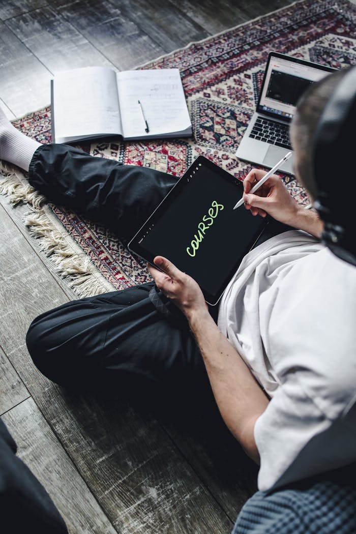 A young adult using a tablet to write courses while seated on a patterned rug indoors.