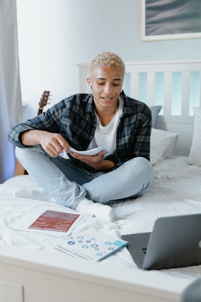 A young man studies online with a laptop and notes, sitting comfortably on his bed.