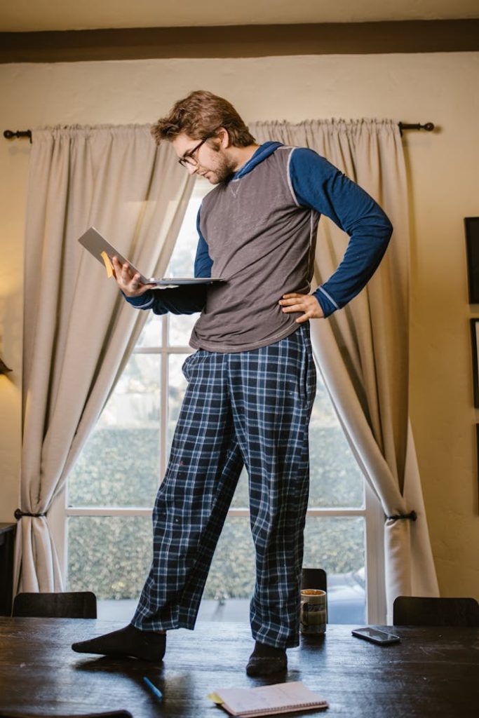 Man in pajama pants standing on table using laptop for study in cozy home setting.